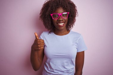 Genç afrikalı afro kadın izole pembe arka plan üzerinde t-shirt gözlük giyen el ile jest kadar mutlu başparmak yapıyor. Başarı göstererek kameraya bakan ifadeyi onaylama.