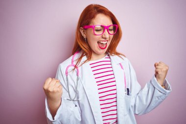 Genç kızıl doktor kadın izole pembe arka plan üzerinde duran stetoskop kullanarak çok mutlu ve heyecanlı kolları kaldırdı ile kazanan jest yapıyor, gülümseyerek ve başarı için çığlık. Kutlama konsepti.