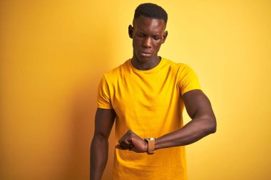 Genç afro-Amerikan adam izole sarı arka plan üzerinde duran rahat t-shirt giyen kol saati zaman kontrol, rahat ve kendinden emin