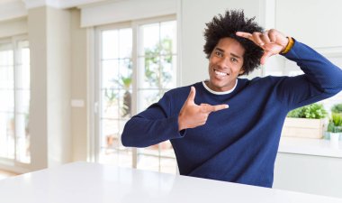 Genç afro-amerikan adam evde oturan rahat kazak giyen mutlu yüzü ile elleri ve parmakları ile çerçeve yapma gülümseyerek. Yaratıcılık ve fotoğrafçılık konsepti.