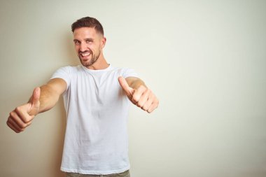 Genç yakışıklı adam izole arka plan üzerinde rahat beyaz t-shirt giyen el ile olumlu jest yapıyor onaylayan, gülümseyen kadar başparmak ve başarı için mutlu. Kazanan jest.