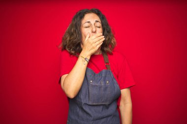 Orta yaş yaşlı kadın kırmızı izole arka plan üzerinde önlük üniforma giyen el ile ağız kapsayan yorgun esneme sıkılmış. Huzursuz ve uykululuk.