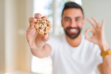 Yakışıklı İspanyol adam çikolatalı kurabiye yiyor parmaklar, mükemmel sembolü ile ok işareti yapıyor