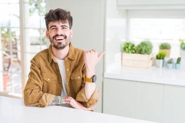 Beyaz masanın üzerinde oturan rahat ceket giyen genç adam mutlu yüz görünümlü ve başparmak ile yan işaret gülümseyerek.