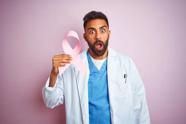 Young indian doctor man holding cancer ribbon standing over isolated pink background scared in shock with a surprise face, afraid and excited with fear expression