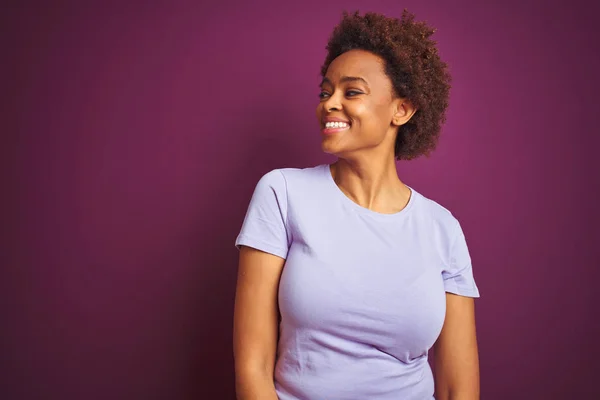 Young beautiful african american woman with afro hair over isolated purple background looking away to side with smile on face, natural expression. Laughing confident.