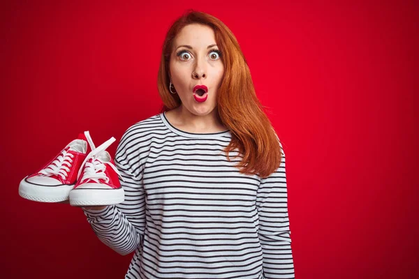 Jovem Mulher Ruiva Bonita Segurando Tênis Sobre Fundo Isolado Vermelho — Fotografia de Stock