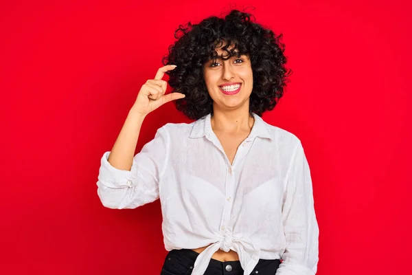 年轻的阿拉伯妇女 卷曲的头发 穿着白色休闲衬衫在孤立的红色背景微笑和自信的手势与手做小尺寸的标志 手指看和相机 测量概念 — 图库照片