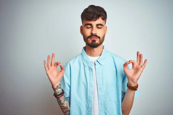 Young man with tattoo wearing blue shirt standing over isolated white background relax and smiling with eyes closed doing meditation gesture with fingers. Yoga concept.