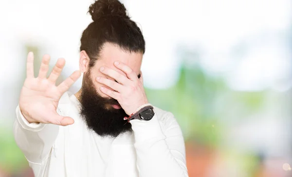 Young man with long hair and beard wearing sporty sweatshirt covering eyes with hands and doing stop gesture with sad and fear expression. Embarrassed and negative concept.