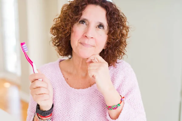资深妇女拿着粉红色牙刷在牙科诊所严肃面对思考问题 想法很困惑 — 图库照片
