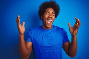 Afro saçlı afro saçlı adam izole mavi arka plan üzerinde duran t-shirt giyen silah kaldırdı ve kapalı gözleri heyecan çığlık ile başarı için deli ve deli kutluyor. Kazanan kavramı
