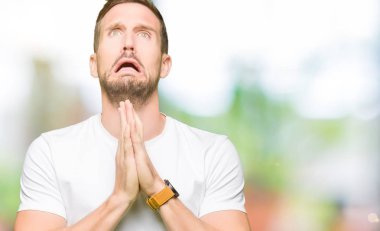 Yakışıklı adam yalvarıyor ve umut ifade yüzünde çok duygusal ve endişe ile birlikte elleriyle dua casual beyaz t-shirt giyiyor. Bağışlanmak. Din kavramı.