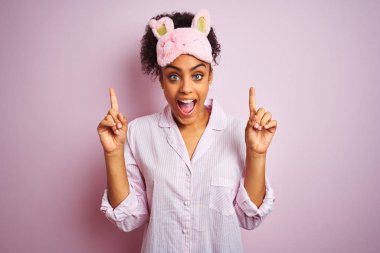 Genç afro-amerikan kadın izole pembe arka plan üzerinde pijama ve maske giyen şaşırmış ve şaşırttı ve parmakları ile işaret ve kolları kaldırdı.