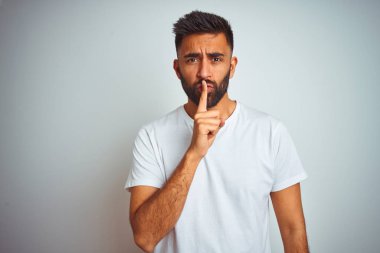İzole beyaz arka plan üzerinde duran t-shirt giyen genç hintli adam dudaklarında parmak ile sessiz olmak isteyen. Sessizlik ve gizli kavram.