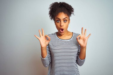 Izole beyaz arka plan üzerinde duran lacivert çizgili t-shirt giyen Afro-amerikan kadın sürpriz görünümlü ve parmakları ile ok onay sembolü yapıyor şok. Çılgın ifade