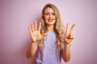 Genç güzel kadın pembe izole arka plan üzerinde duran mor t-shirt giyen gösteren ve emin ve mutlu gülümserken parmakları ile yedi numara işaret.