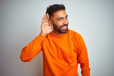Izole beyaz arka plan üzerinde turuncu kazak giyen genç hintli adam söylenti ya da dedikodu bir işitme dinlerken kulak üzerinde el ile gülümseyerek. Sağırlık kavramı.