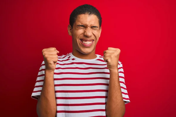 年轻的英俊阿拉伯男子穿着条纹T恤 在孤立的红色背景兴奋的成功与手臂和眼睛闭着庆祝胜利微笑 优胜者概念 — 图库照片