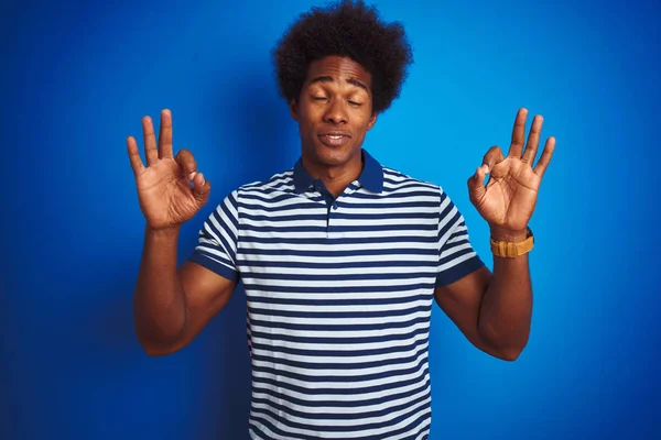 African american man with afro hair wearing striped polo standing over isolated blue background relax and smiling with eyes closed doing meditation gesture with fingers. Yoga concept.