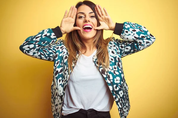 Genç güzel kadın sarı izole arka plan üzerinde rahat ceket giyen gülümseyen neşeli oynarken yüzleri gösteren elleri ile bir boo gözetleme. Şaşırttı ve çıktı