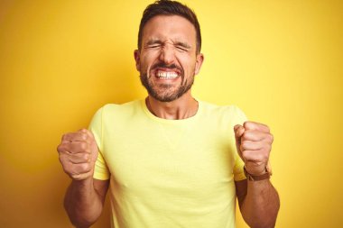 Genç yakışıklı adam sarı izole arka plan üzerinde rahat sarı t-shirt giyen kollarını kaldırdı ve gözleri gülümseyerek zafer kutlayan kapalı başarı için heyecanlı. Kazanan kavramı.