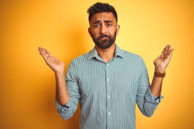 Yeşil çizgili gömlek giyen genç hintli adam izole sarı arka plan üzerinde duran habersiz ve kolları ve elleri ile karışık ifade kaldırdı. Şüphe kavramı.