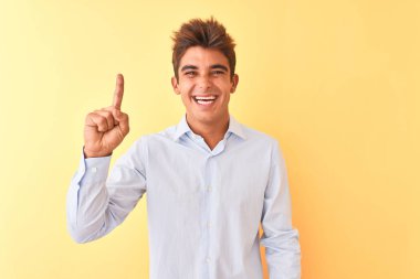 Genç yakışıklı işadamı izole sarı arka plan üzerinde zarif gömlek giyen gösteren ve kendinden emin ve mutlu gülümserken parmak sayısı bir ile işaret.