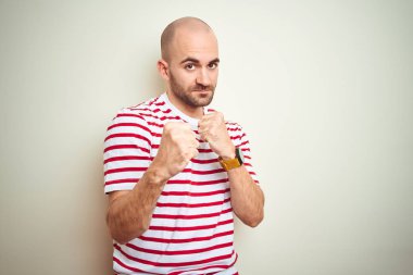 Beyaz izole arka plan üzerinde rahat çizgili kırmızı t-shirt giyen sakallı genç kel adam yumruk savunma jesti ile mücadele etmeye hazır, kızgın ve üzgün yüz, sorun korkuyor