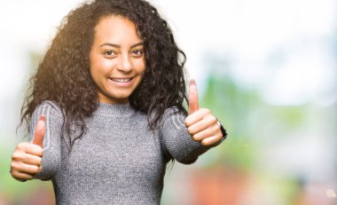 Kıvırcık saçlı genç güzel kız el ile olumlu jest yapıyor onaylayan, başparmak kadar gülümseyerek ve başarı için mutlu. Kameraya bakmak, kazanan jest.