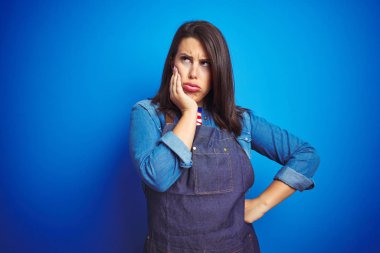 Genç güzel iş kadını mavi izole arka plan üzerinde mağaza üniforma önlük giyen yorgun görünümlü ve çapraz kolları ile depresyon sorunları ile sıkılmış düşünme.