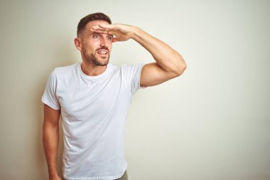 Üzerinde rahat beyaz t-shirt giyen genç yakışıklı adam arka plan çok mutlu ve gülümseyen uzakta el ile üzerine kafa seyir izole. Arama kavramı.