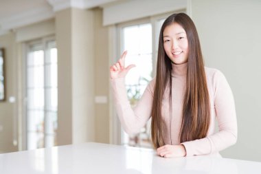 Güzel Asyalı kadın beyaz masada rahat kazak giyen gülümseyerek ve emin el ile el yaparken boyutu işareti parmakları ile yaparken ve kamera. Ölçü kavramı.