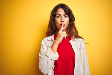 Kırmızı t-shirt giyen genç güzel kadın ve sarı izole arka plan üzerinde çizgili gömlek dudaklarında parmak ile sessiz olmak isteyen. Sessizlik ve gizli kavram.