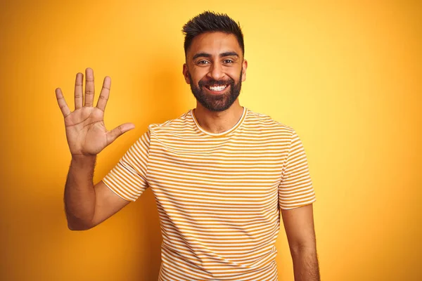 孤立した黄色の背景の上に立つTシャツを着た若いインド人男性は 自信を持って幸せに微笑みながら 5番の指で見せ 指を上げている — ストック写真