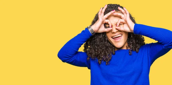 Young beautiful woman with curly hair wearing winter sweater doing ok gesture like binoculars sticking tongue out, eyes looking through fingers. Crazy expression.