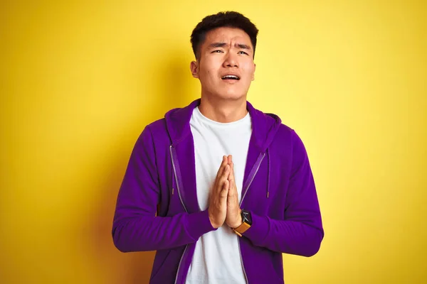 紫色のスウェットシャツを着たアジアの若い中国人男性は 孤立した黄色の背景の上に立って 非常に感情的で心配した顔に希望の表情と手をつないで祈ります 許しを請う宗教概念 — ストック写真