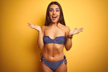 Tatilde bikini giyen genç güzel kadın izole sarı arka plan üzerinde duran deli kutluyor ve kollarını kaldırdı ve açık gözleri heyecan çığlık ile başarı için hayran. Kazanan kavramı