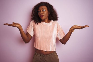 Genç afro-amerikan kadın izole pembe arka plan üzerinde duran t-shirt giyen habersiz ve kolları ve elleri ile karışık ifade kaldırdı. Şüphe kavramı.