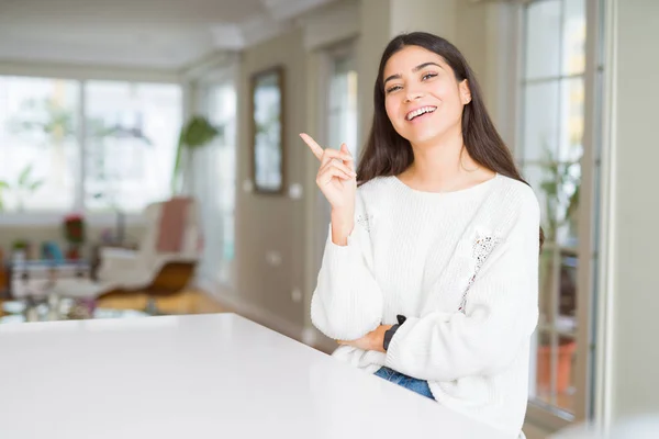 年轻美丽的女人在家里白色的桌子上 脸上挂着灿烂的笑容 用手和手指指着一旁看着镜头 — 图库照片