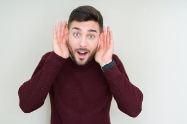 İzole arka plan üzerinde bir kazak giyen genç yakışıklı adam kulak jestiki elini duymak için çalışıyorum, dedikodu için meraklı. İşitme sorunu, sağır