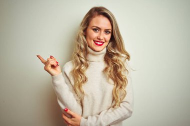 Yüzünde büyük bir gülümseme ile izole beyaz arka plan üzerinde kış boğazlı kazak giyen güzel kadın, kameraya bakarak yan el parmağı ile işaret.