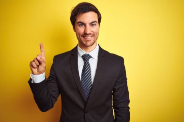 Takım elbise giyen ve kravat giyen genç yakışıklı işadamı izole sarı arka plan üzerinde duran gösteren ve kendinden emin ve mutlu gülümserken parmak sayısı bir işaret.