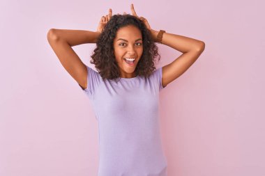 Genç Brezilyalı kadın izole pembe arka plan üzerinde duran t-shirt giyen tavşan kulakları gibi baş parmakları ile komik ve deli poz, neşeli gülümseyen