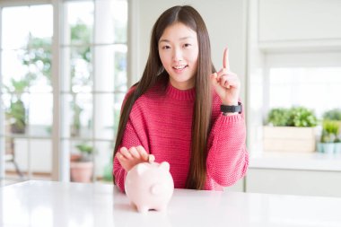 Güzel Asyalı kadın bir fikir ya da mutlu yüz, bir numara ile işaret parmağı ile şaşırttı piggy çanta içine bir sikke koyarak