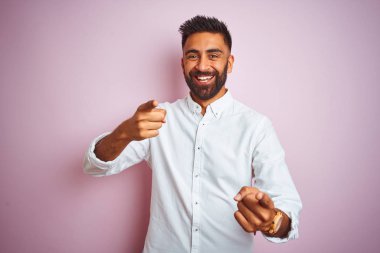 Genç Hintli işadamı mutlu ve komik yüzü ile kameraya parmaklarını gösteren izole pembe arka plan üzerinde duran zarif gömlek giyiyor. İyi enerji ve titreşimler.