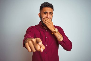 Kırmızı zarif gömlek giyen genç hintli adam izole gri arka plan üzerinde duran size gülüyor, ağız üzerinde el ile kameraya parmak işaret, utanç ifade