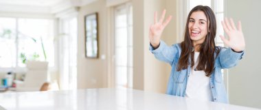 Geniş açı resim gösterme ve parmakları ile işaret evde beyaz masaya oturan güzel genç kadının kendinden emin ve mutlu gülümseyerek süre sekiz numara.