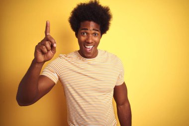 Afro saçlı Amerikalı adam çizgili t-shirt izole sarı arka plan üzerinde başarılı bir fikir ile parmak işaret üzerinde duran giyiyor. Çıktı ve mutlu. Bir numara.