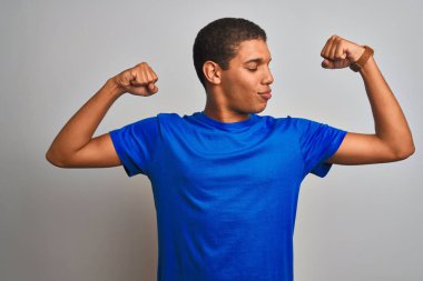 Genç yakışıklı Arap adam mavi t-shirt giyen izole beyaz arka plan üzerinde duran kol kasları gururla gülümseyen gösteren. Fitness konsepti.